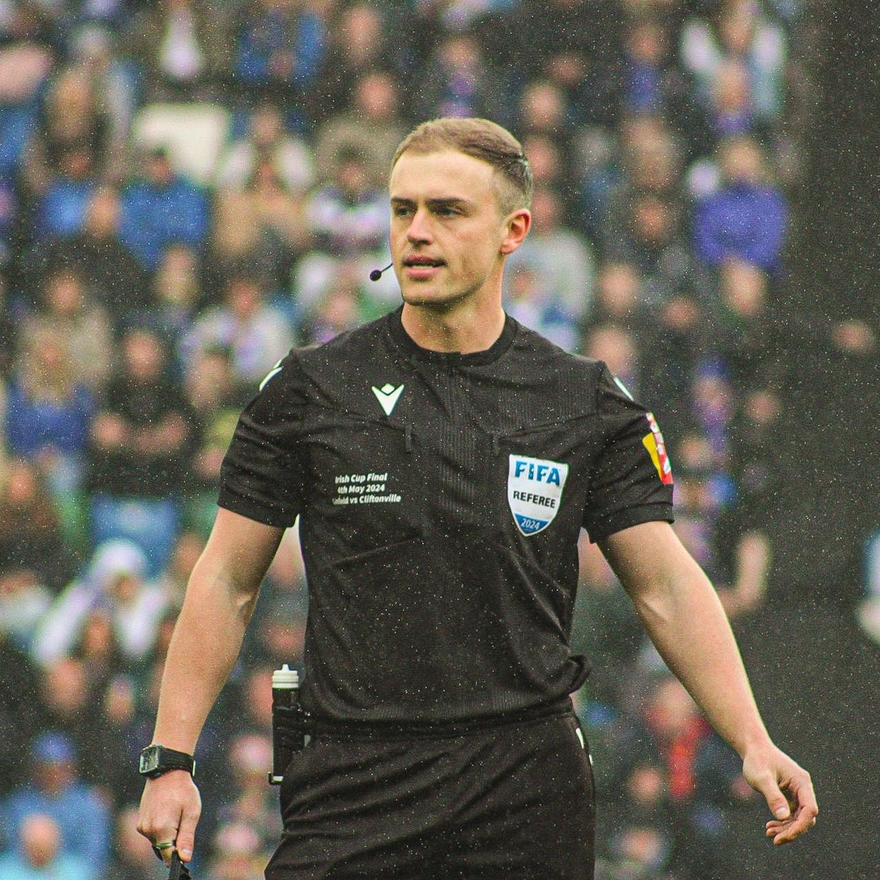 A referee in a black uniform with a FIFA badge stands on a football field, looking focused. A blurred crowd is visible in the background, enhancing the action setting.